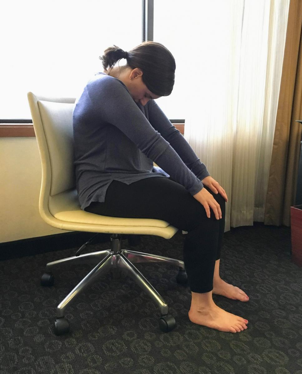 Woman sitting in chair with her back hunched and head dropping forward, hands on her knees with arms straigh