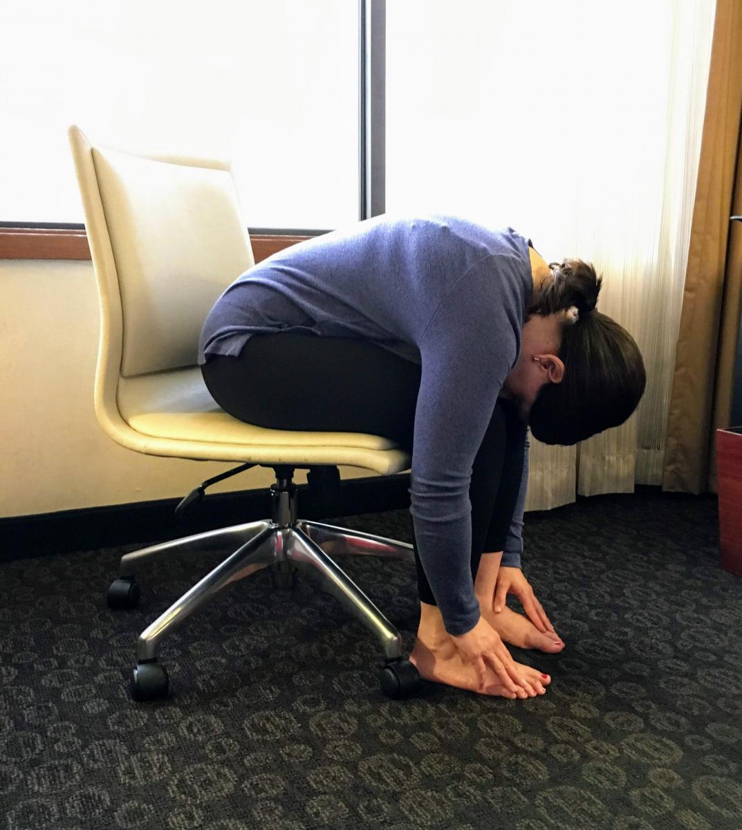 Woman sitting in chair with her torso folded flat over her lap and hands reaching down to her feet