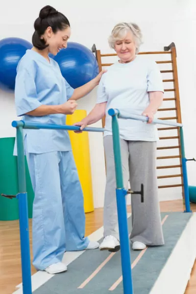 older woman using parallel bars to walk with the help of a physical therapist.