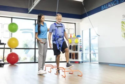 Physical therapist helping patient wearing a harness step over obstacles.